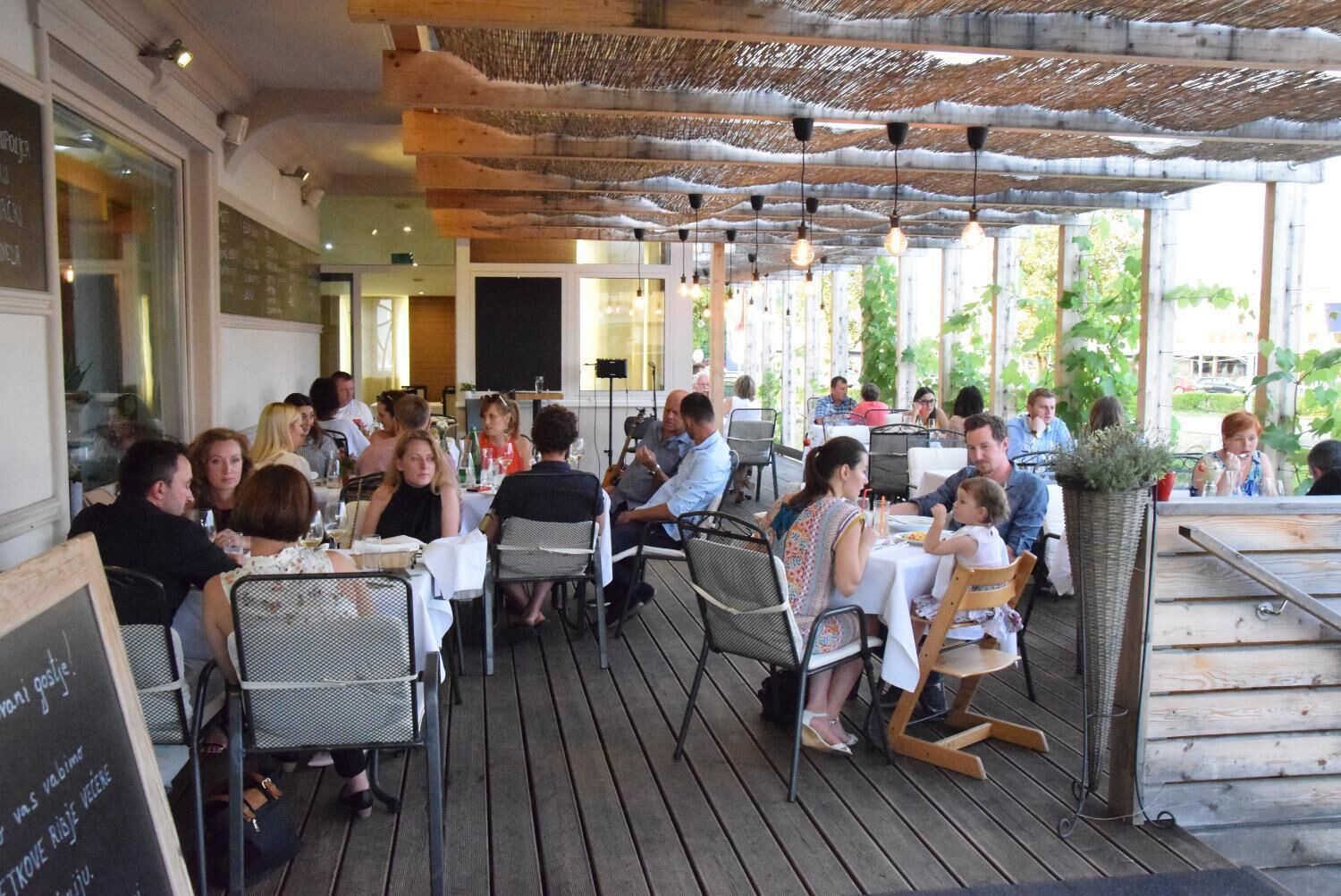 An outdoor dining area with wooden tables and chairs, covered by a thatched roof.