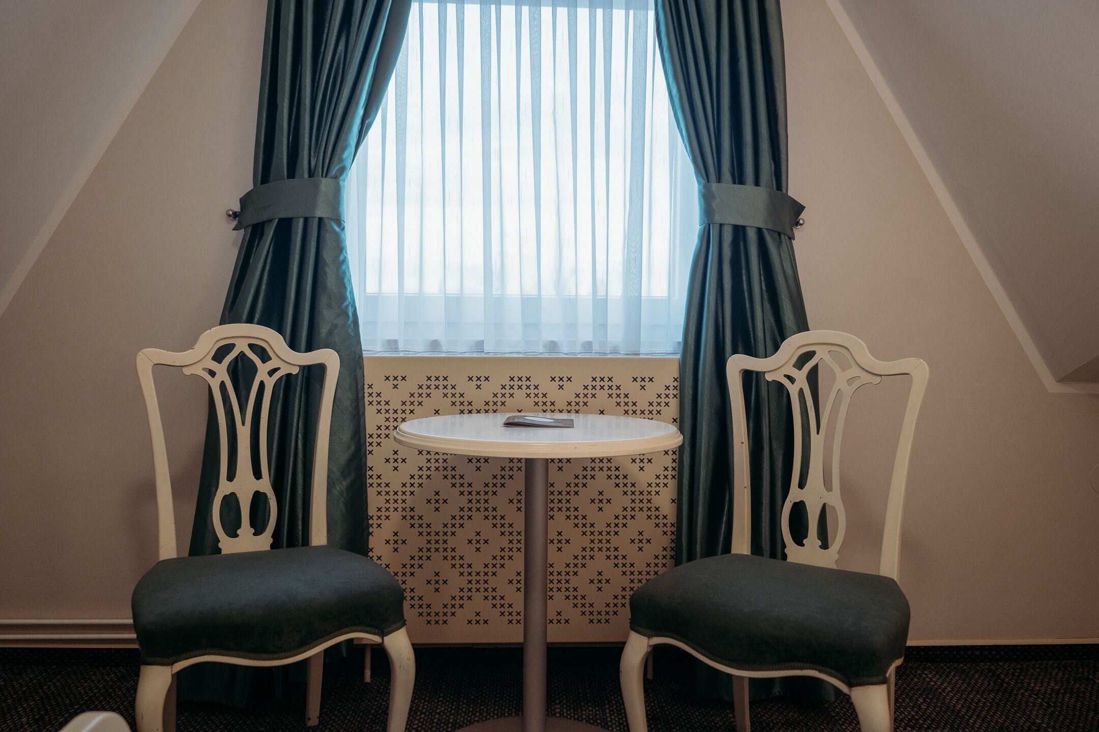 A small room with two white chairs, a round table, and a window with blue curtains.