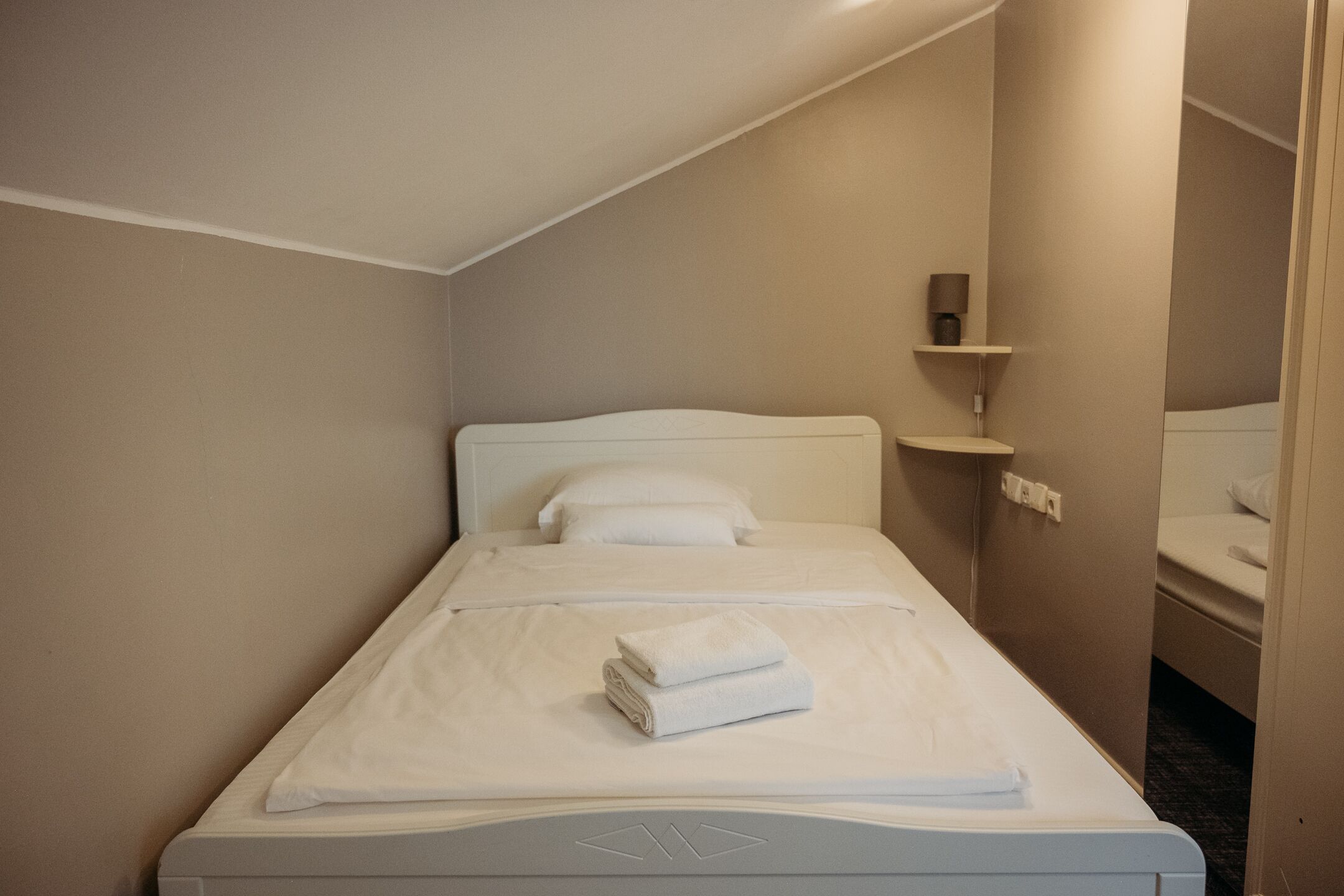 Una cama bien hecha con sábanas blancas y una toalla doblada, una lámpara de noche y una pequeña estantería con una lámpara.
