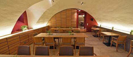 A restaurant with a high, arched ceiling and wooden panel walls. Tables are set with chairs and potted plants.