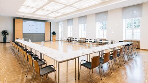 Un salón de conferencias con una larga mesa, sillas, una pantalla de proyector y ventanas amplias.