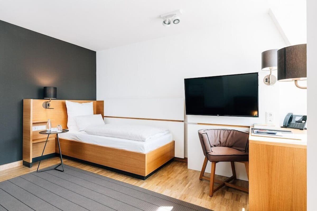 A modern hotel room with a bed, desk, chair, TV, and a small table with a lamp.