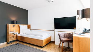 A modern hotel room with a wooden bed, a desk with a chair, a flat-screen TV, and a small table with a lamp.