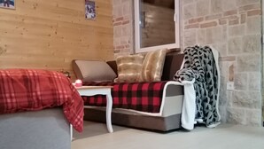 Una habitación acogedora con techo de madera, pared de piedra, ventana, sofá con manta y una pequeña mesa.