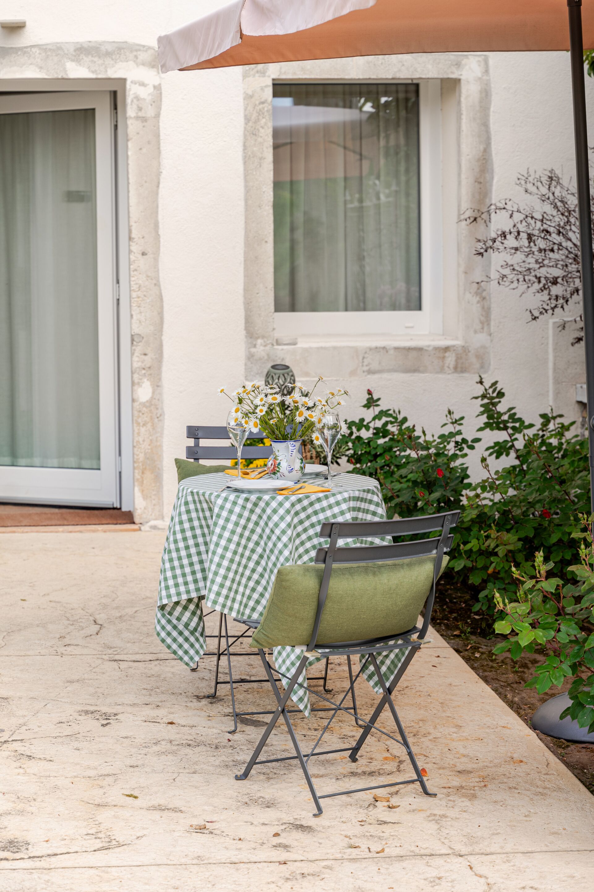 An outdoor dining area with a checkered tablecloth, two chairs, a vase with flowers, and a window.