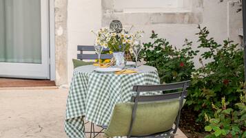 An outdoor dining area with a checkered tablecloth, two chairs, a vase with flowers, and a window.