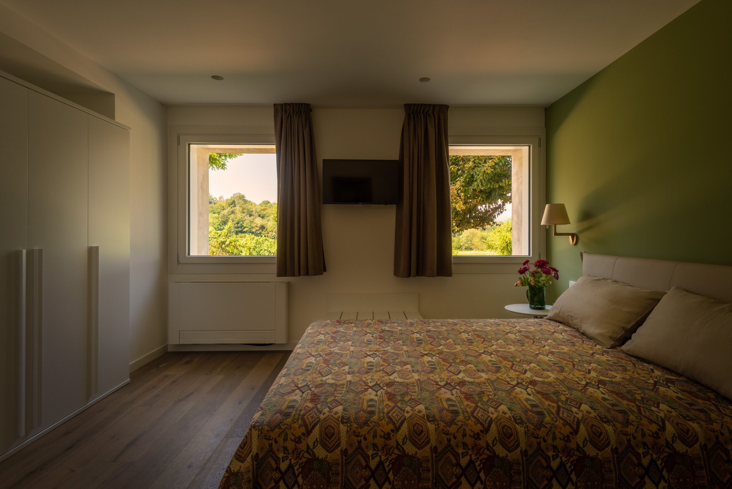 A bedroom with a bed, a TV mounted on the wall, a window with curtains, a bedside table with a lamp, and a radiator.
