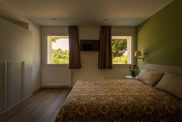 A bedroom with a bed, a TV mounted on the wall, a window with curtains, a bedside table with a lamp, and a radiator.