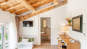 Una habitación con techo de madera, un sofá blanco con cojines verdes, una mesa de comedor de madera con sillas, un televisor montado en la pared y un baño visible a través de una puerta abierta.