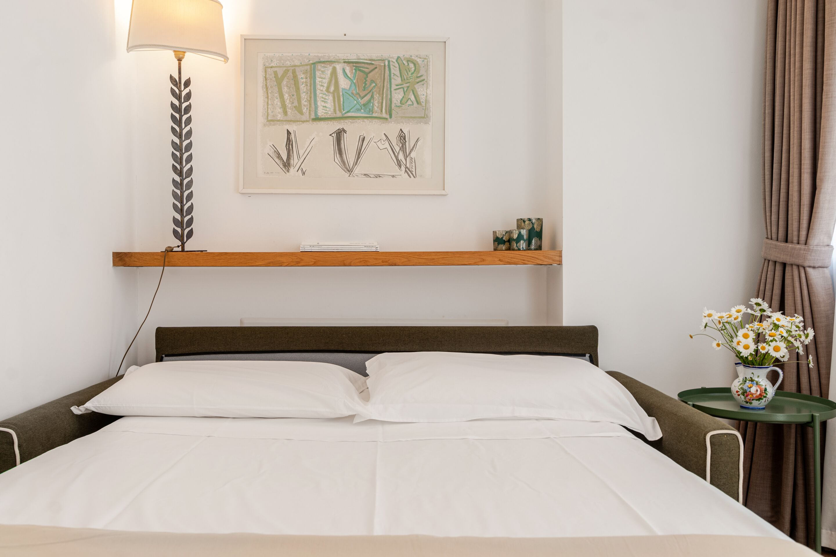 A neatly made bed with white linens, a bedside lamp, a framed abstract artwork, a small table with a vase of flowers, and a wooden shelf with decorative items.
