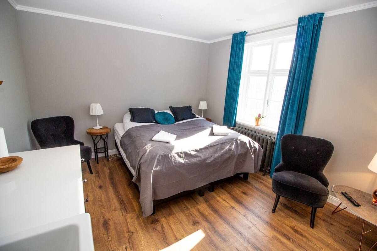 Une chambre à coucher avec un lit, deux chaises, une petite table, un radiateur et une fenêtre avec des rideaux.