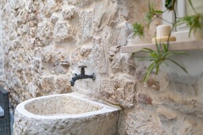 Un lavamanos de piedra con un grifo frente a una pared de piedra texturizada, un estante pequeño con velas y una planta en maceta.