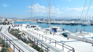 Un port de plaisance avec des bateaux amarrés, une vue sur la ville en arrière-plan et une promenade piétonne bordée de garde-corps.
