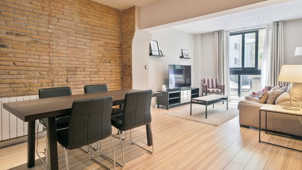Een moderne woonkamer met een bakstenen muur, een eethoek met een tafel en stoelen, een bank, een televisie en een raam met gordijnen.