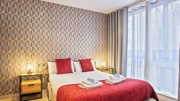 Ein ordentlich bezogenes Bett mit roter Decke, zwei Nachttische mit Lampen, eine Wand mit geometrischem Muster und ein großes Fenster mit durchsichtigen Vorhängen.