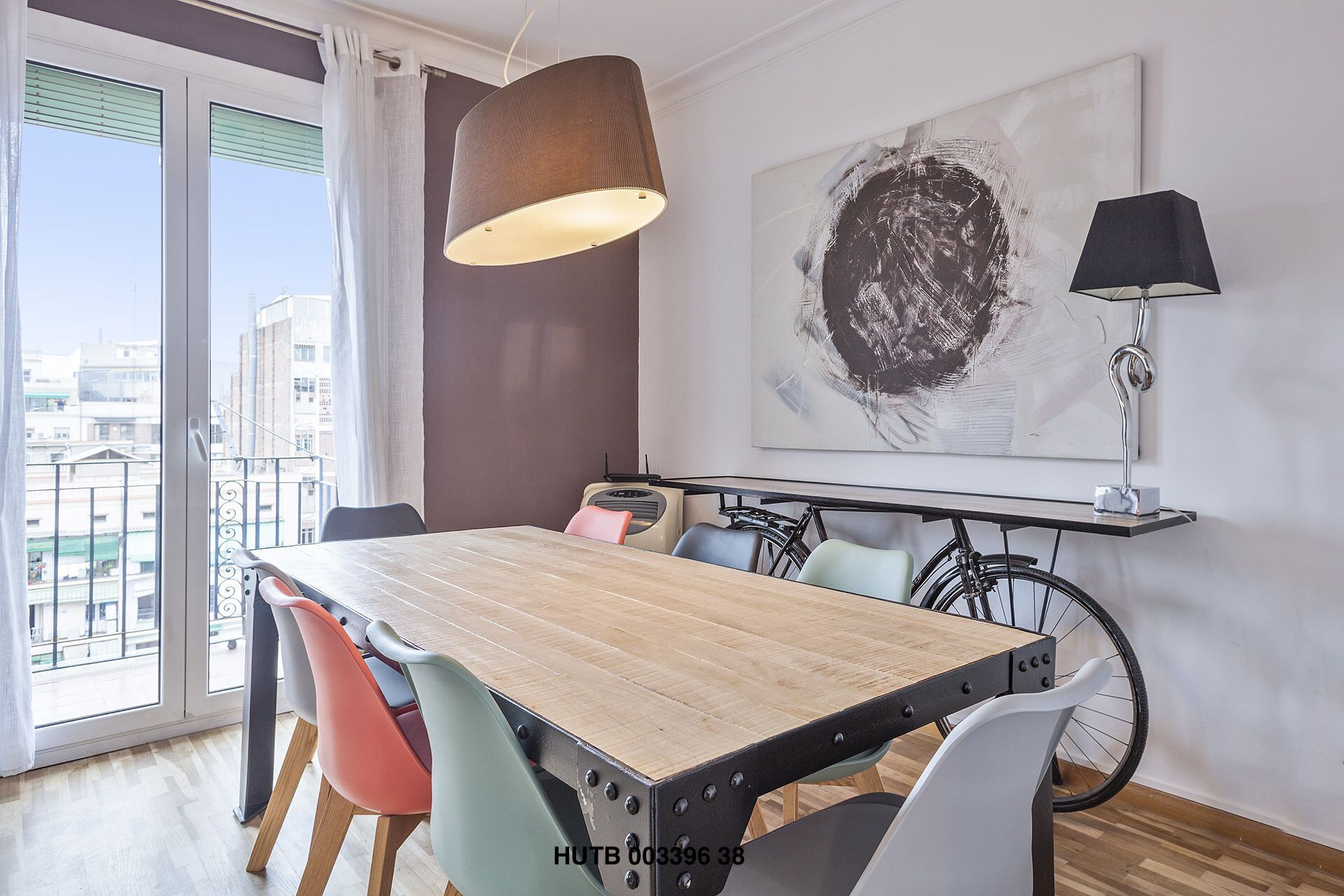 A dining area with a wooden table, colorful chairs, a hanging light fixture, a bicycle, and a large abstract painting on the wall.