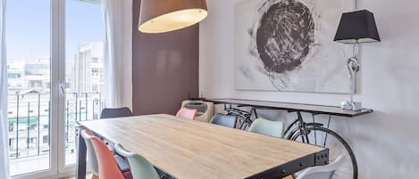 A dining area with a wooden table, colorful chairs, a hanging light fixture, a bicycle, and a large abstract painting on the wall.