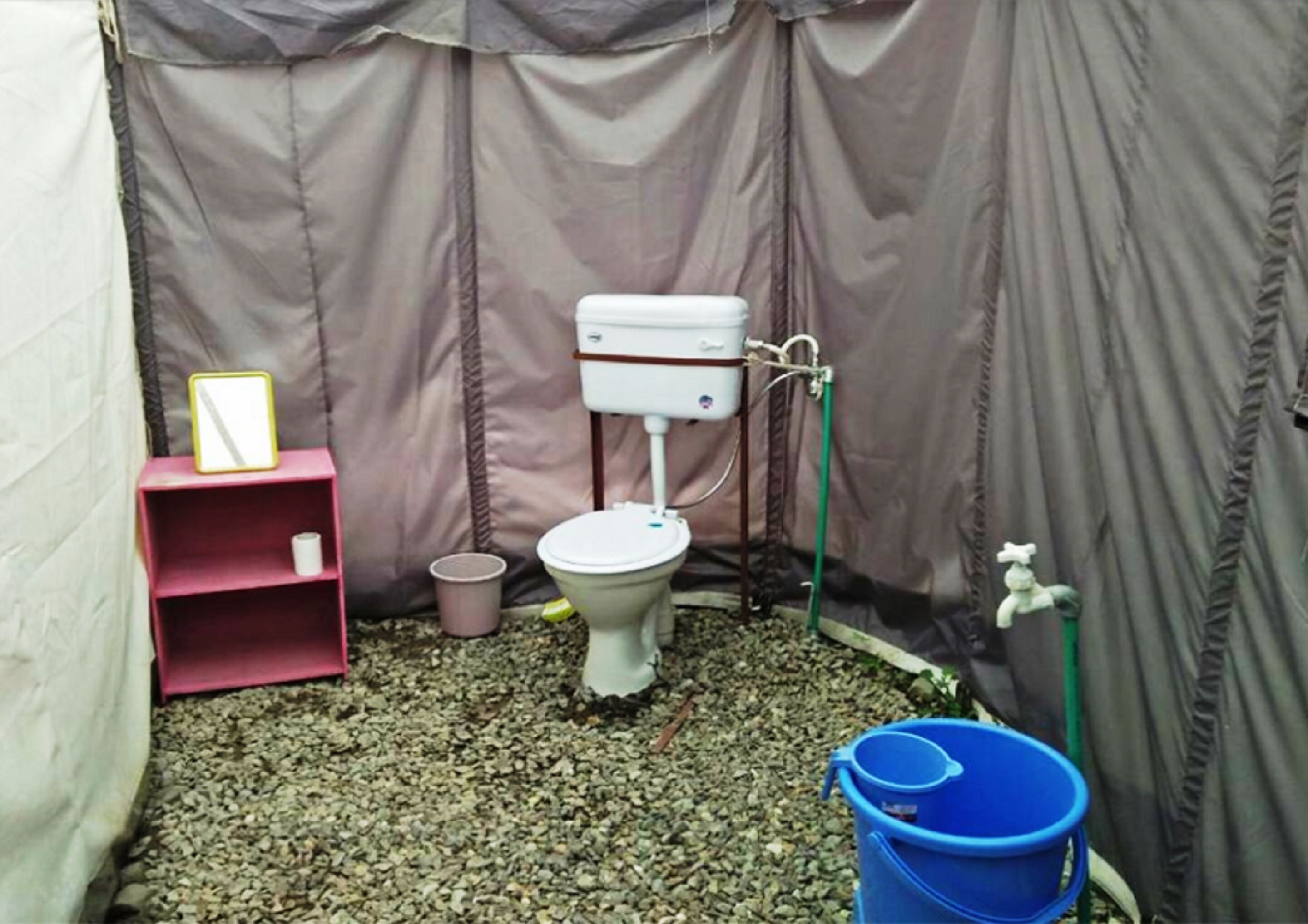 A small, enclosed bathroom with a toilet, a pink shelf with a mirror, a blue bucket, and a green hose.