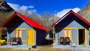 Deux petites cabanes, chacune composée d’une seule pièce, avec des toits rouges et des détails jaunes, et une chaise placée à l’extérieur de chacune.