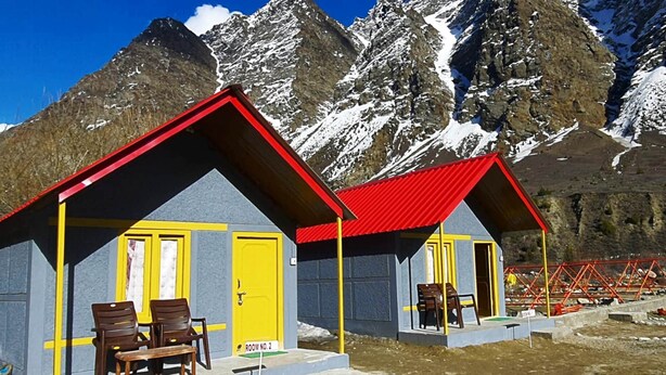 Deux petits bâtiments d’un seul étage, avec des toits rouges et des portes jaunes, situés dans une région montagneuse, avec des sommets enneigés en arrière-plan.