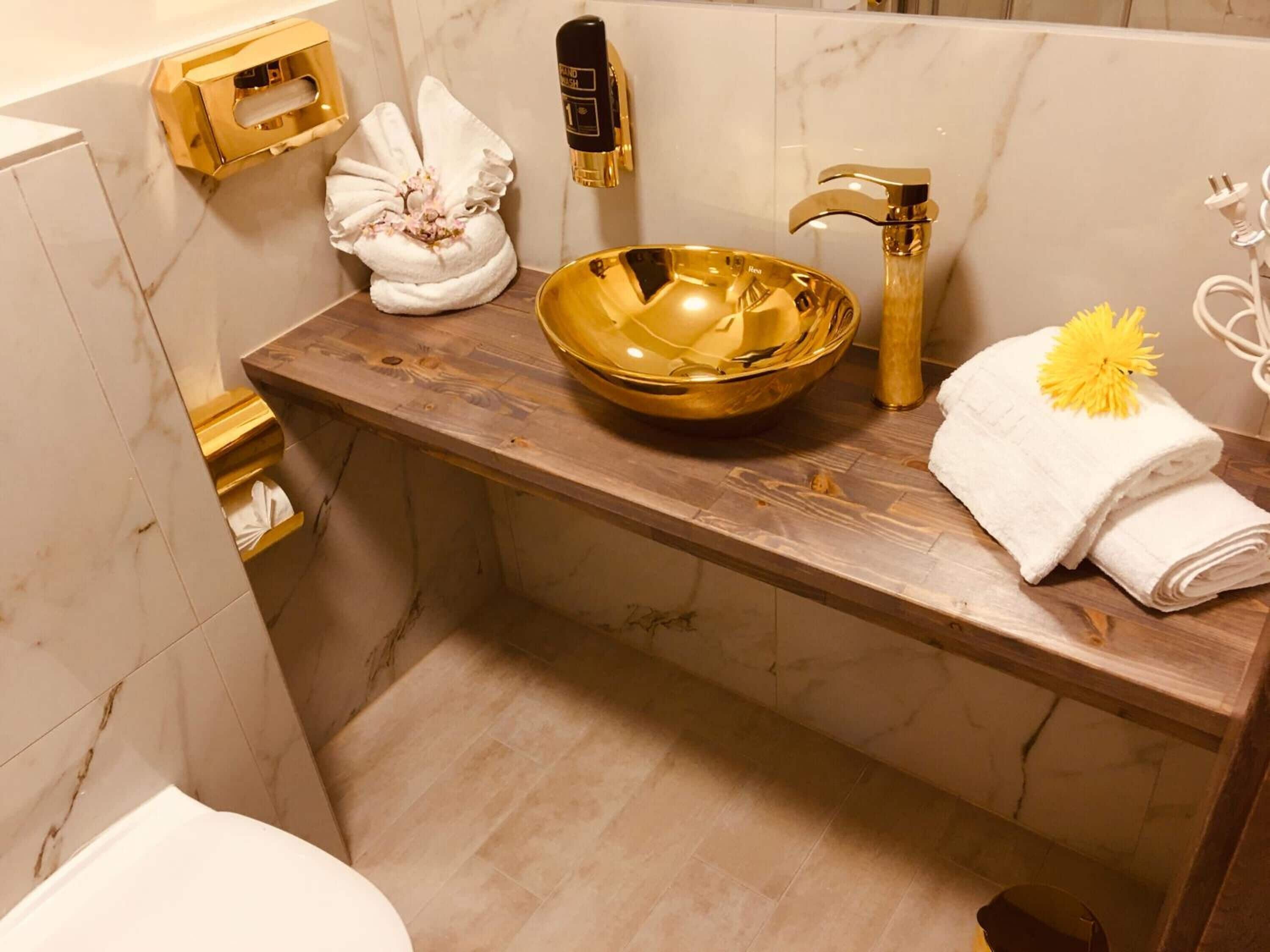 Un baño con pared de mármol, tocador de madera, lavabo dorado y toalla con un estampado de flor amarilla.