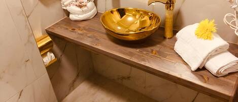 Un baño con pared de mármol, tocador de madera, lavabo dorado y toalla con un estampado de flor amarilla.