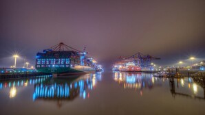 A large cargo ship docked at a port with cranes and a calm water surface reflecting the ship and surrounding lights.