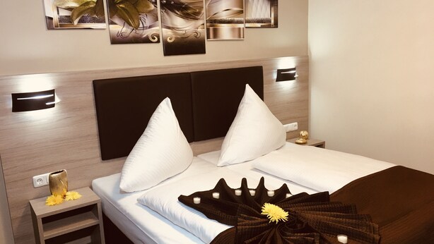 A modern hotel room with a large bed, two white pillows, a dark brown throw, and a small bedside table with a lamp and a decorative item.