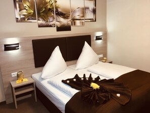 A modern hotel room with a large bed, two white pillows, a dark brown throw, and a small bedside table with a lamp and a decorative item. - Hotel Glinde (Glinde)