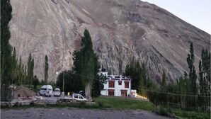 A mountainous landscape with a building featuring a white facade and red accents, surrounded by trees and parked vehicles. - TIH Namra Village Hotel - Temisgam (Leh)
