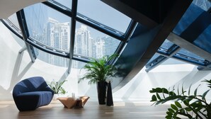 Un interior moderno con techo curvo, un amplio ventanal con vistas a la ciudad, una silla azul y una planta en maceta.