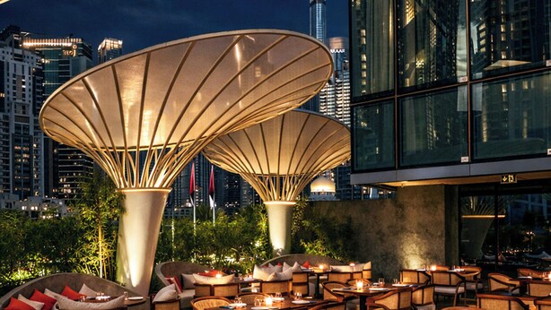 Un espace extérieur aménagé pour les repas, avec plusieurs tables et chaises, de grands parasols et, en arrière-plan, une vue sur les bâtiments de la ville.