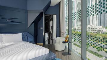 A modern hotel room with a large bed, a seating area, and a view of the cityscape through large windows.
