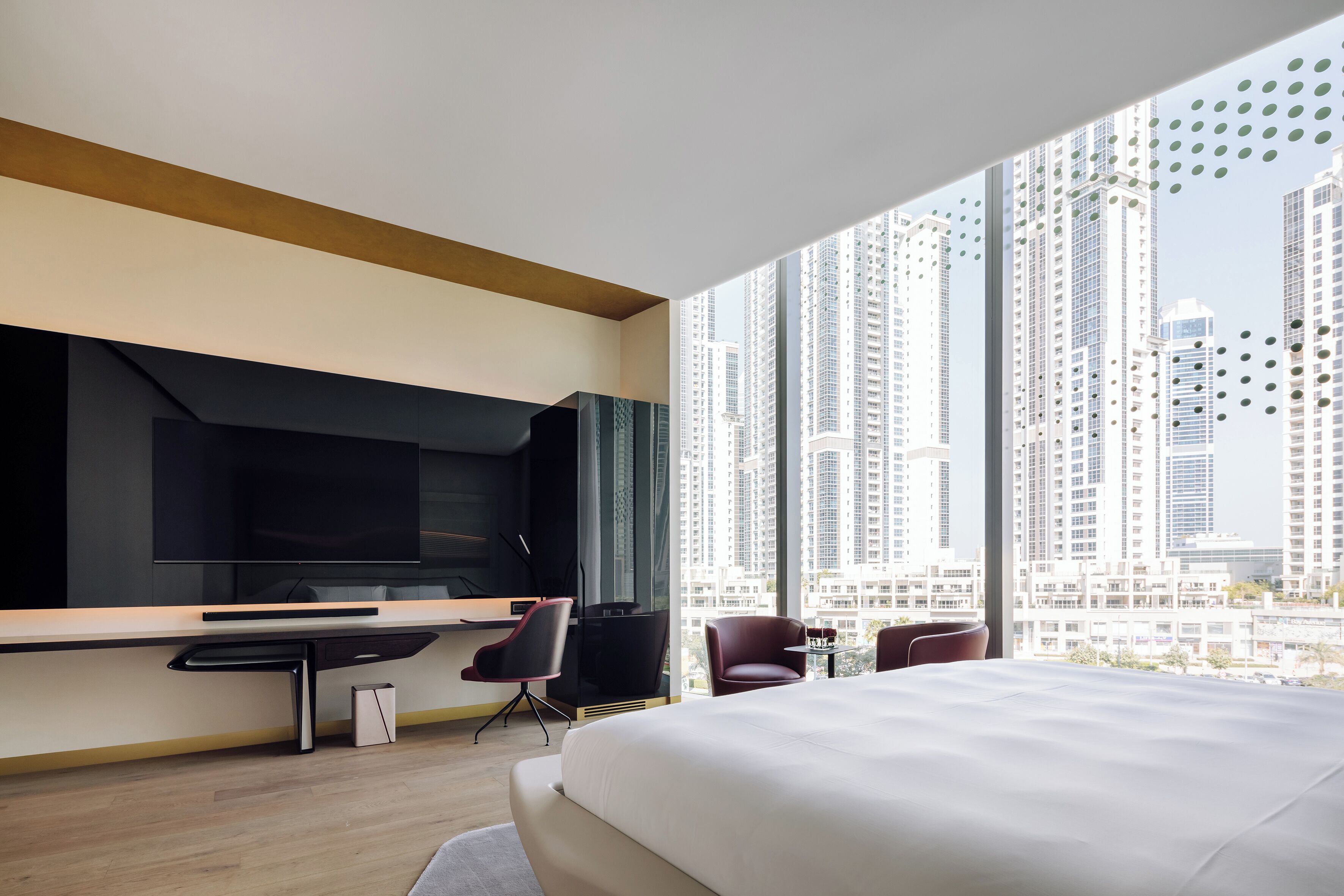 A modern hotel room with a large bed, a desk with a flat-screen TV, and a view of tall buildings.
