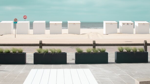 A beachside walkway with white planters and a row of white structures, possibly beach huts, along the shore. - State-of-the-art studio with frontal sea view (Koksijde-Bad)