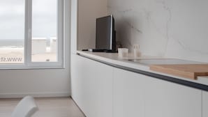 A modern kitchen with a large window overlooking a beach, a flat-screen TV, and a wooden cutting board on the counter. - State-of-the-art studio with frontal sea view (Koksijde-Bad)