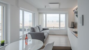 A modern, well-lit room with a glass dining table, a grey sofa, and a large window offering a view of the beach. - State-of-the-art studio with frontal sea view (Koksijde-Bad)