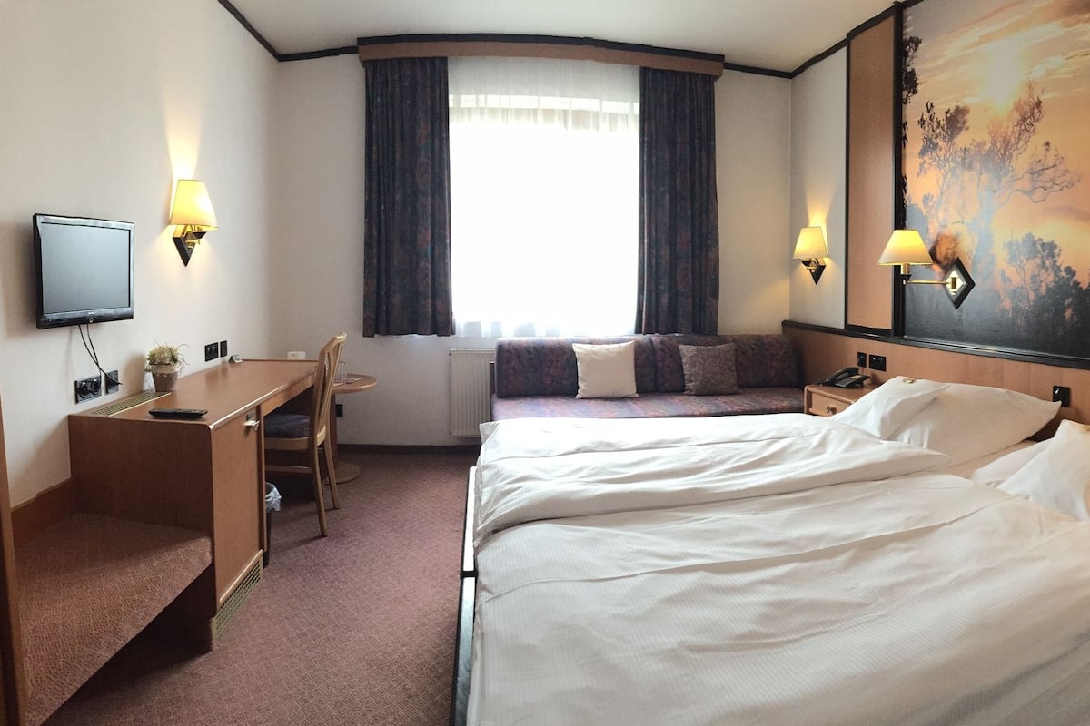 A hotel room with a bed, a desk, a chair, a television, and a painting of a sunset.