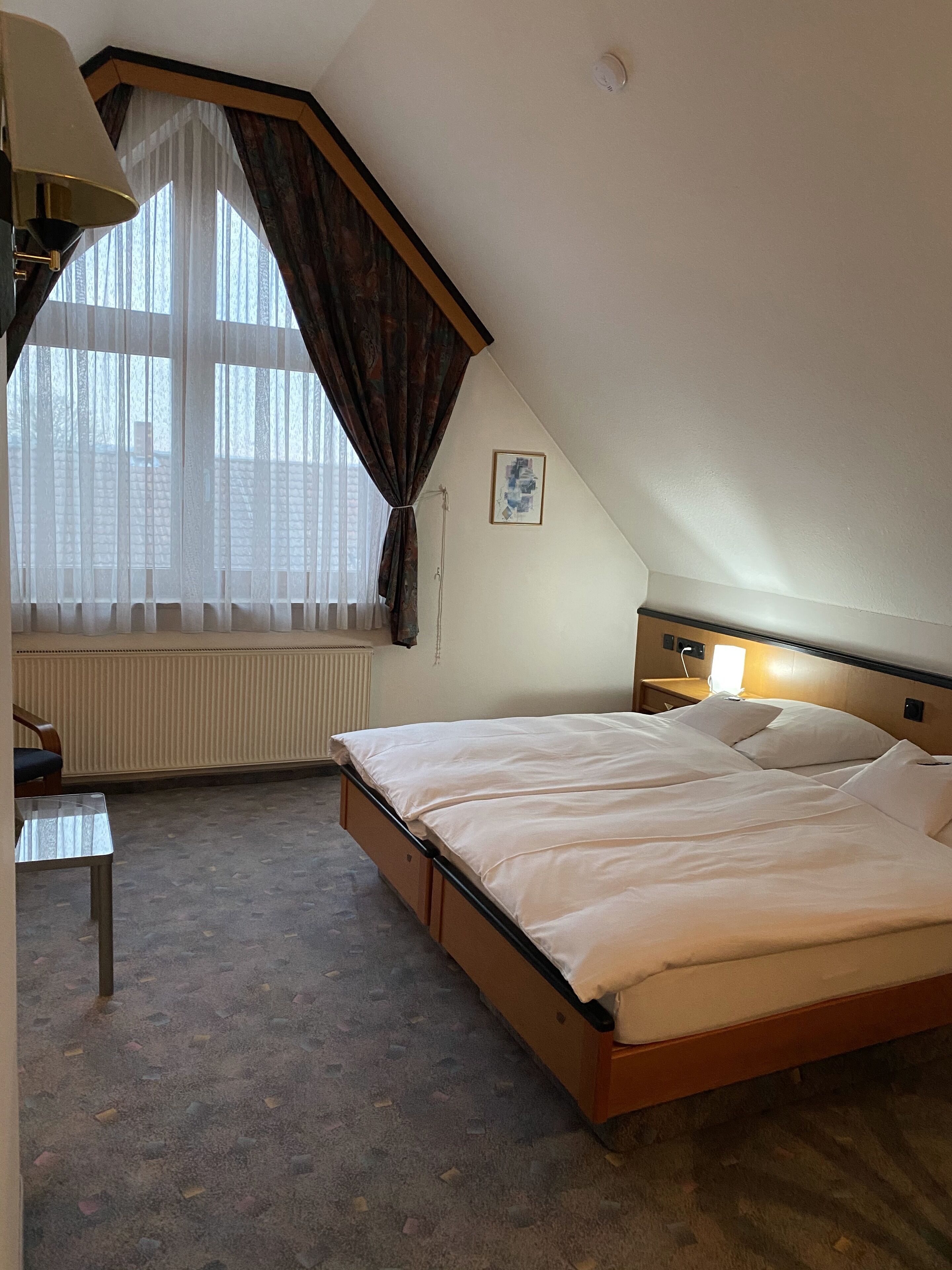 A hotel room with a bed, a desk, a lamp, a window with curtains, a radiator, and a picture on the wall.