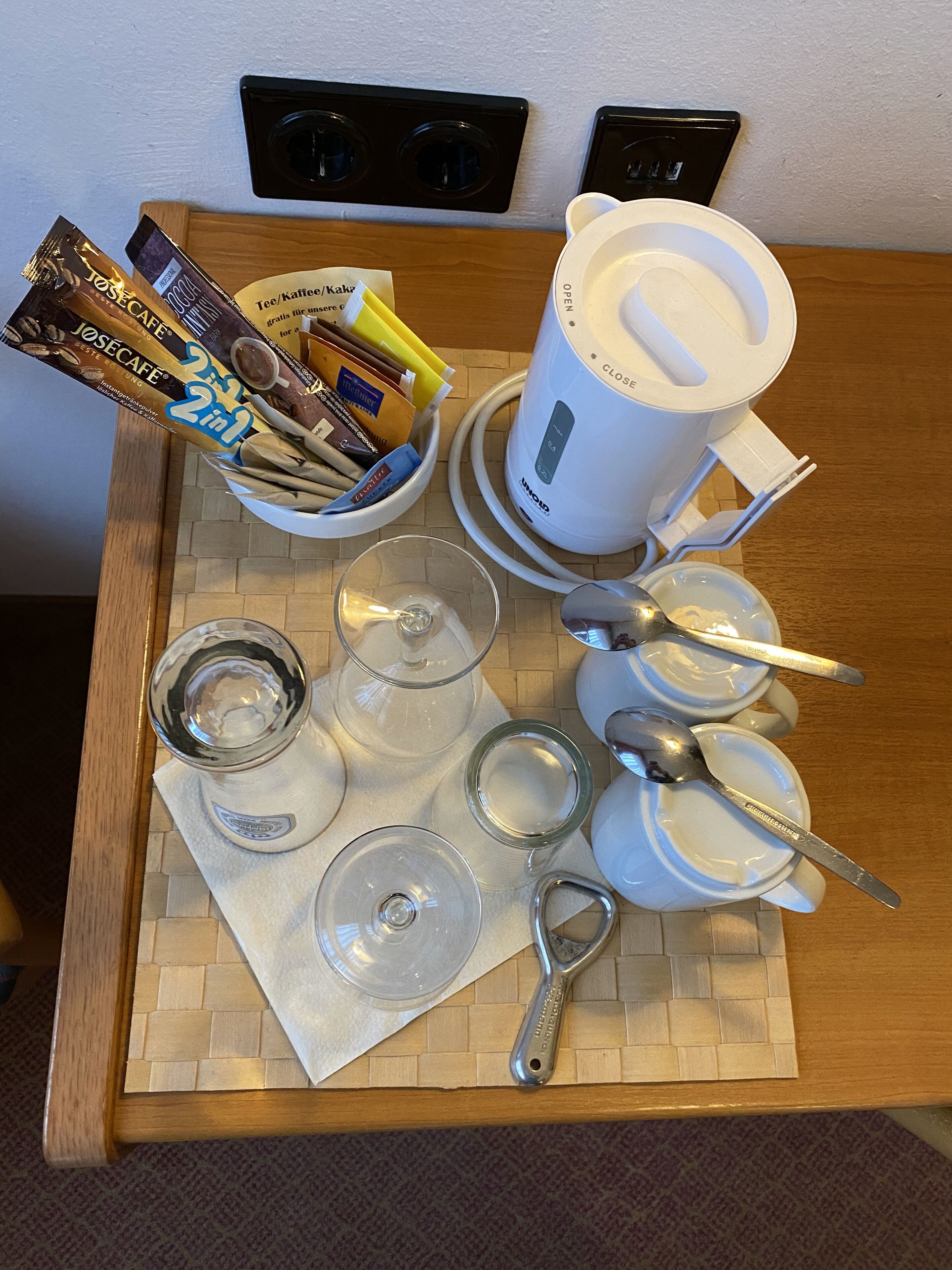Una bandeja de desayuno con cápsulas de café, cafetera, vasos y utensilios sobre una mesa de madera.