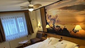 A hotel room with a large bed, two bedside tables, a small round table, a chair, a ceiling fan, and a wall mural. - Hotel Seebach (Grossenseebach)