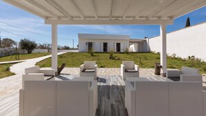 Een overdekt terras met rieten meubels, een modern gebouw op de achtergrond en een heldere lucht.