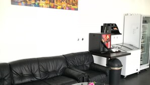 A modern living room with a black leather sofa, a glass coffee table with a pink flower vase, a refrigerator, and a vending machine. - Jura Hotel (Nuremberg)