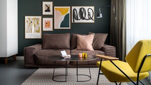 A modern living room with a brown sofa, a yellow armchair, and a round coffee table. There are framed artworks on the wall and a floor lamp. - BodenseeApartments (Friedrichshafen)
