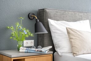 A neatly made bed with a white pillow, a bedside table with a lamp, a glass vase with flowers, and a power outlet. - BodenseeApartments (Friedrichshafen)