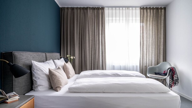 A modern hotel room with a large bed, a bedside table with a lamp, a chair, and a view of the outdoors through sheer curtains. - BodenseeApartments (Friedrichshafen)