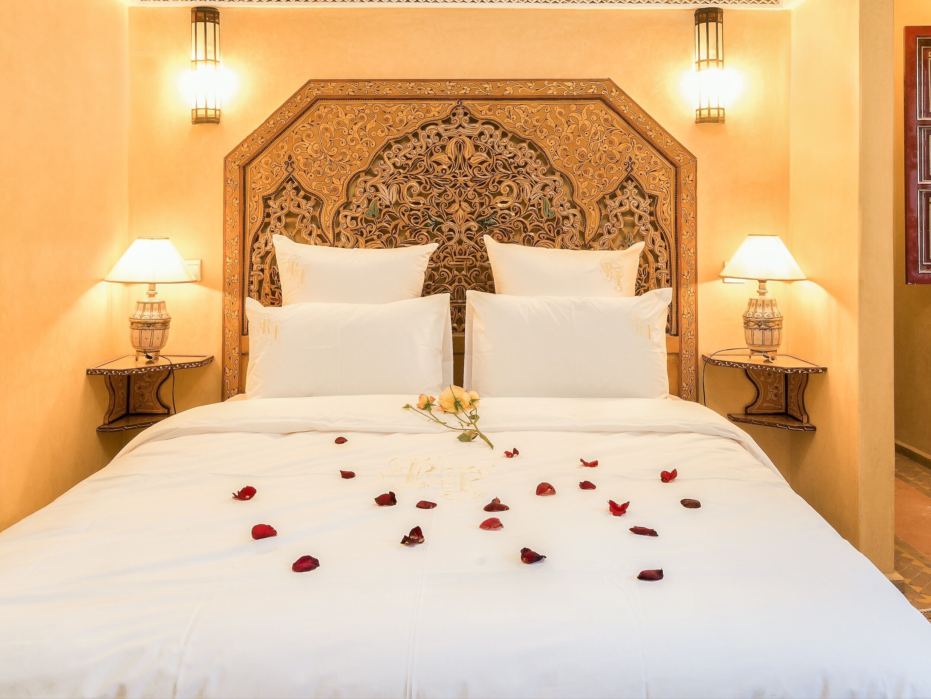A bed with white linens, two pillows, and rose petals scattered on the bed.