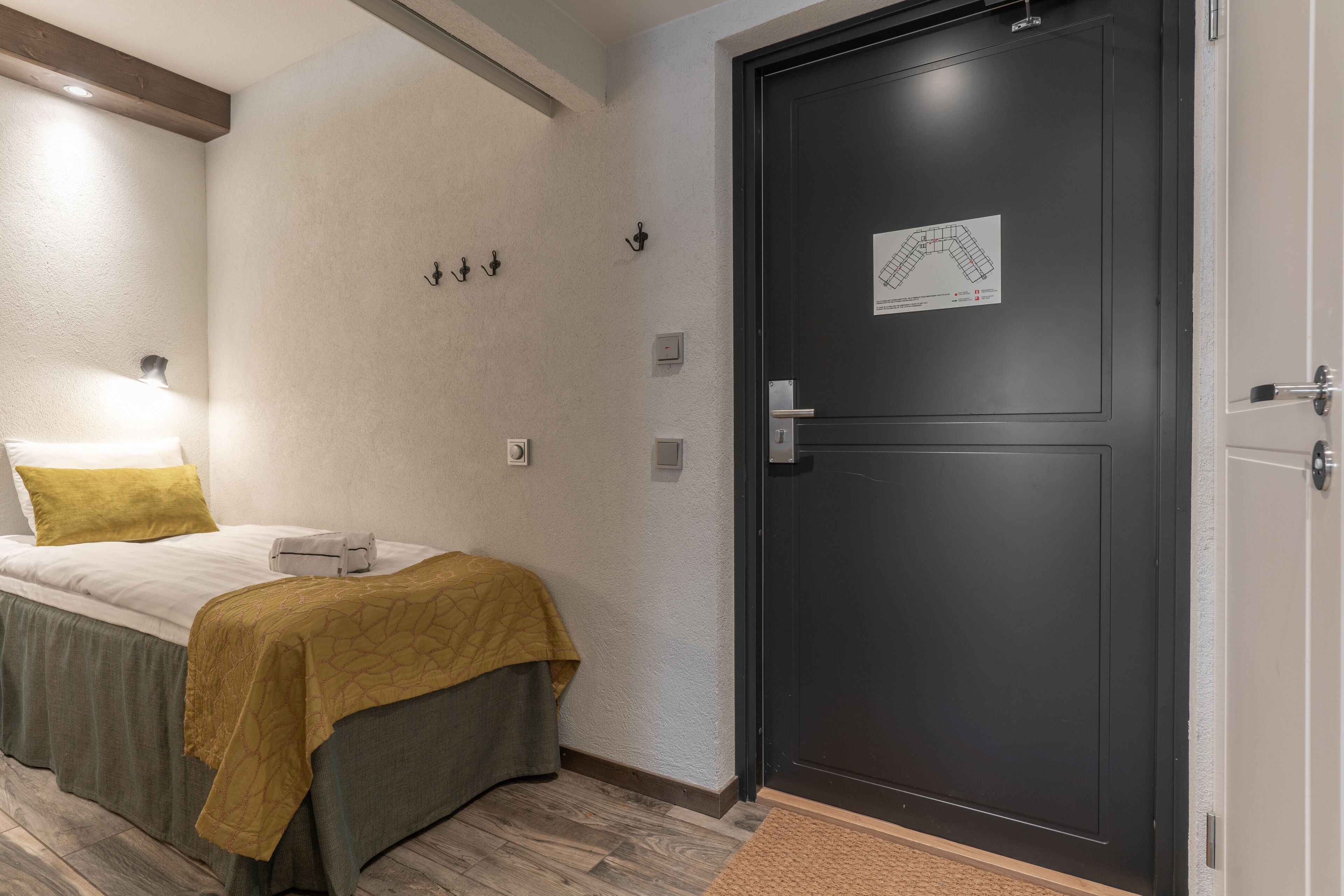 Camera d'albergo con un letto, una porta scura, una lampada fissata al muro e una piccola mappa sulla porta.
