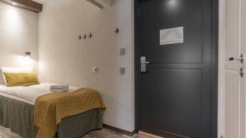 Camera d'albergo con un letto, una porta scura, una lampada fissata al muro e una piccola mappa sulla porta.