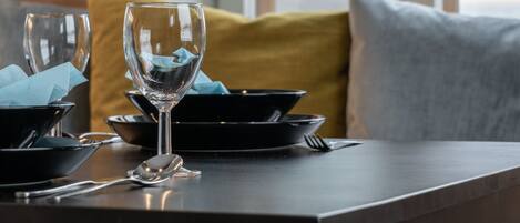 A dining table with a wine glass, black plates, and cutlery. There are yellow and grey cushions on the bench.